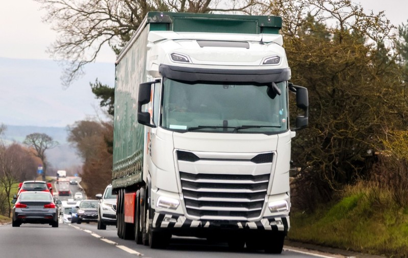 Un camion alb deplasându-se pe o șosea alături de alte autoturisme, într-o zonă de drum mărginită de vegetație.