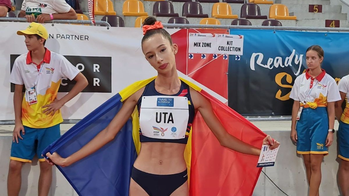 Alexandra Ștefania Uță, în echipament de atletism, cu steagul României pe spate și tribunele pe fundal