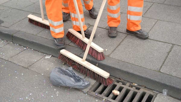 Picioarele unor lucrători de la salubritate purtând pantaloni portocalii reflectorizanți și măturând un trotuar și bordura unei străzi acoperite de resturi și deșeuri.   