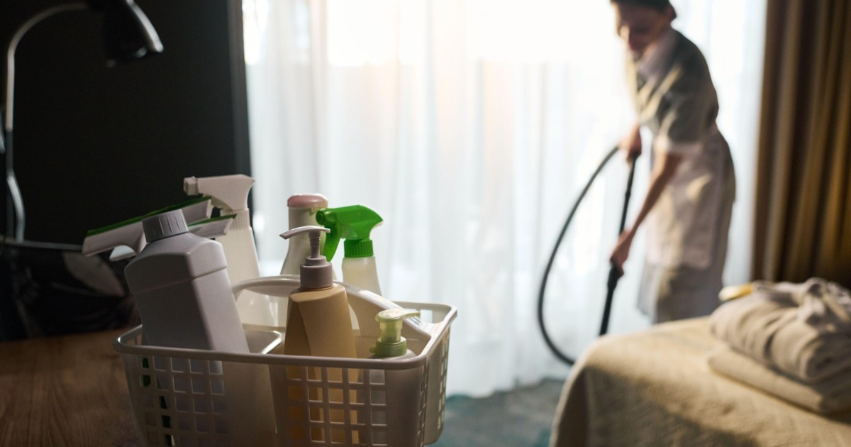 Imaginea prezintă în prim-plan un coș cu produse de curățenie, iar în fundal o cameristă în uniformă care aspiră podeaua unei camere de hotel.