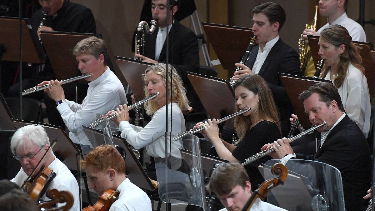 În imagine, o orchestră cântă la Festivalul George Enescu, muzicienii manipulează mai multe instrumente muzicale de suflat și câteva viori