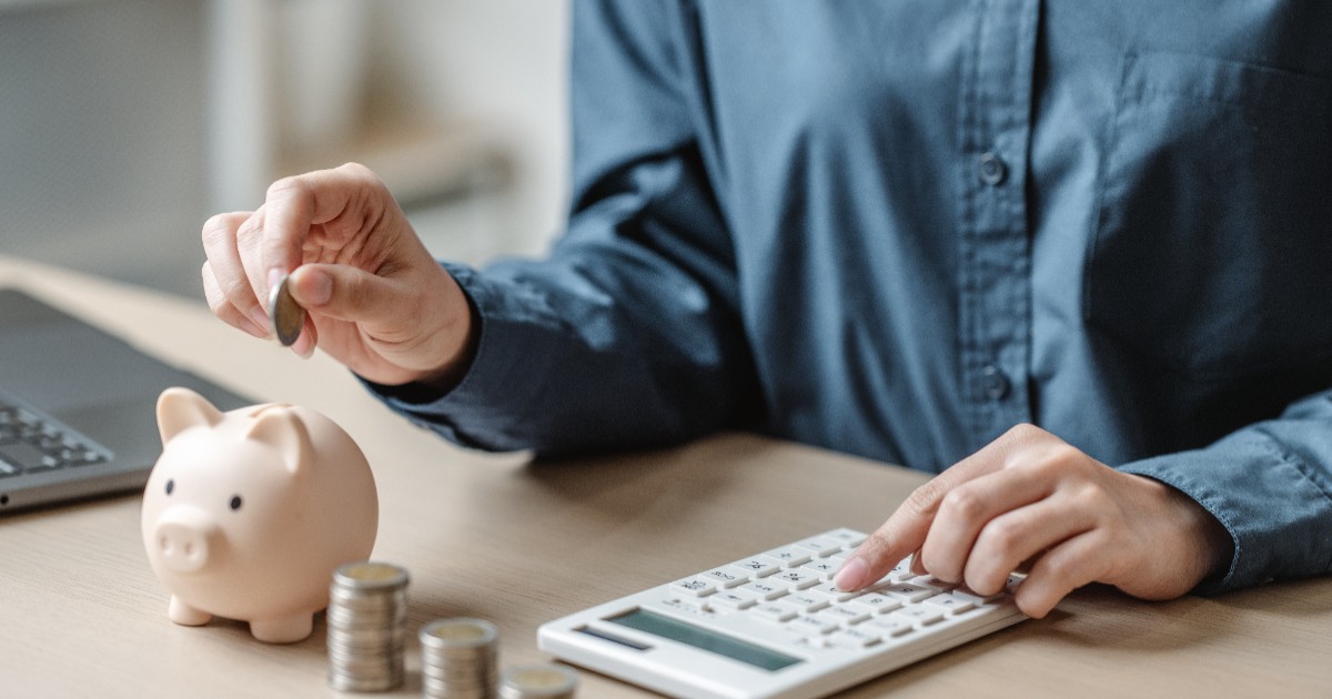 Un bărbat care face calcule pe un calculator, la birou. Pe acesta se află mai multe monede și un porcușor.