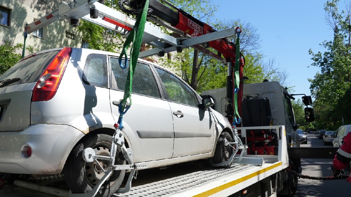 Imaginea arată o mașină argintie, un model de tip hatchback, care este ridicată și transportată pe platforma unei mașini de tractări. Mașina de tractări are un braț hidraulic roșu și verde, cu cabluri și sisteme de prindere specializate pe roți, folosite pentru a ridica și a asigura vehiculul. În fundal se pot vedea clădiri de locuințe și copaci, sugerând că acțiunea se desfășoară într-o zonă urbană, probabil o parcare sau o stradă. Vremea pare a fi însorită.
