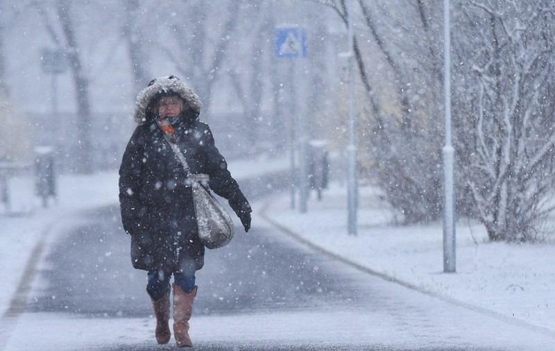 O persoană îmbrăcată cu o geacă groasă cu glugă cu blană mergând pe un trotuar acoperit de zăpadă în timpul unei ninsori abundente.