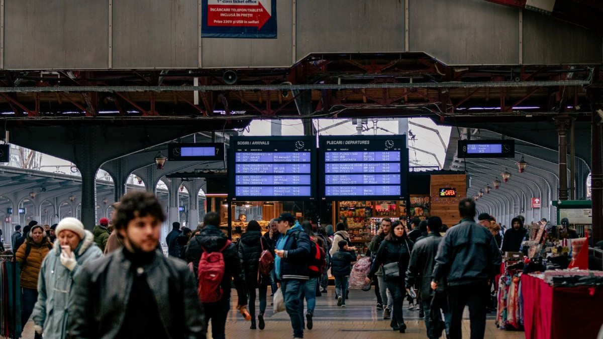 Imagine creativă cu Gara de Nord din București, în prim plan, în blur, mai mulți călători, iar pe fundal panourile de informare cu plecări și sosiri ale trenurilor