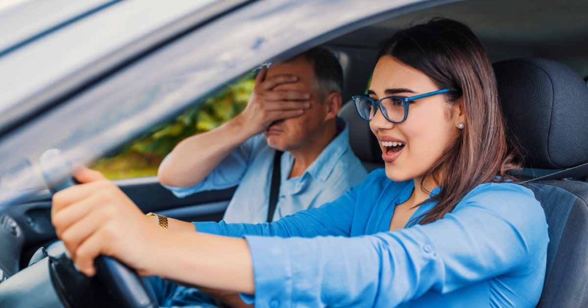 O femeie la volan, în timpul orelor de condus, alături de instructorul auto care își ține mâinile la ochi