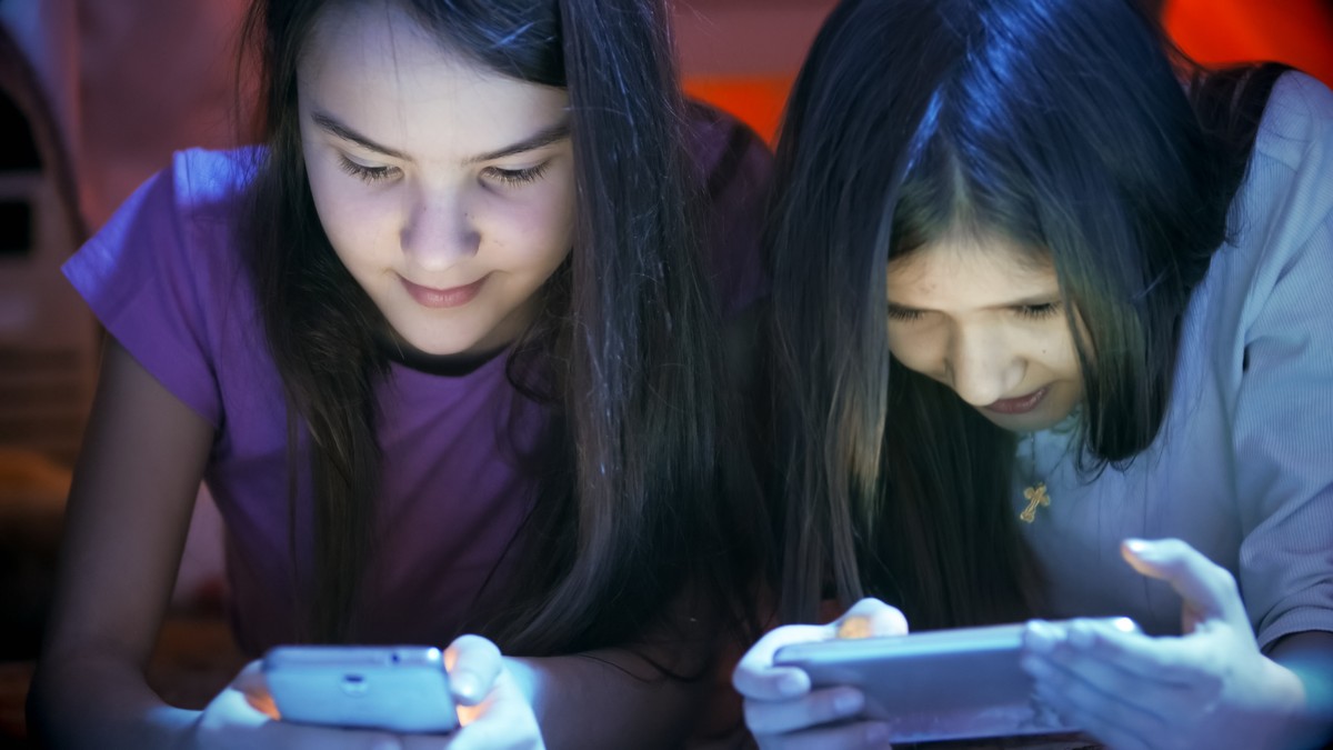 De la ce vârstă îi dai copilului telefon sau laptop conectat la internet: 36% dintre români zic de la 14%, iar 27% de la 10. Două fete care stau pe smartphone-uri, în pat