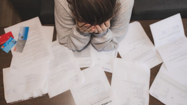 O persoană care își ține capul în mâini deasupra unei mese pline de facturi, documente împrăștiate și carduri bancare, sugerând o stare de stres sau epuizare financiară