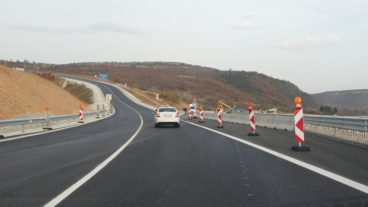 Un autoturism alb care circulă pe o secțiune proaspăt asfaltată de șosea sau autostradă, delimitată de jaloane rutiere și lucrări în desfășurare pe margine.