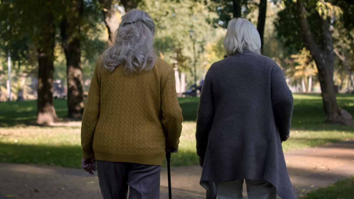 Două femei în vârstă, îmbrăcate în haine de lână și mergând în baston, pe o aless dintr-un parc
