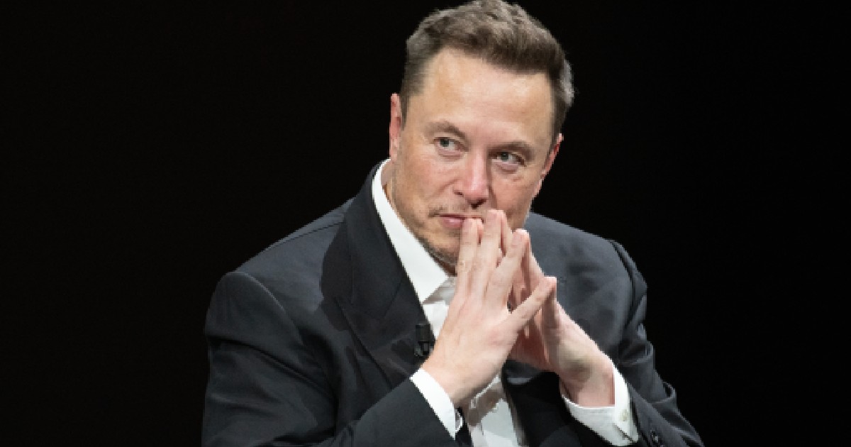 Elon Musk îmbrăcat în costum își ține mâinile în formă de triunghi