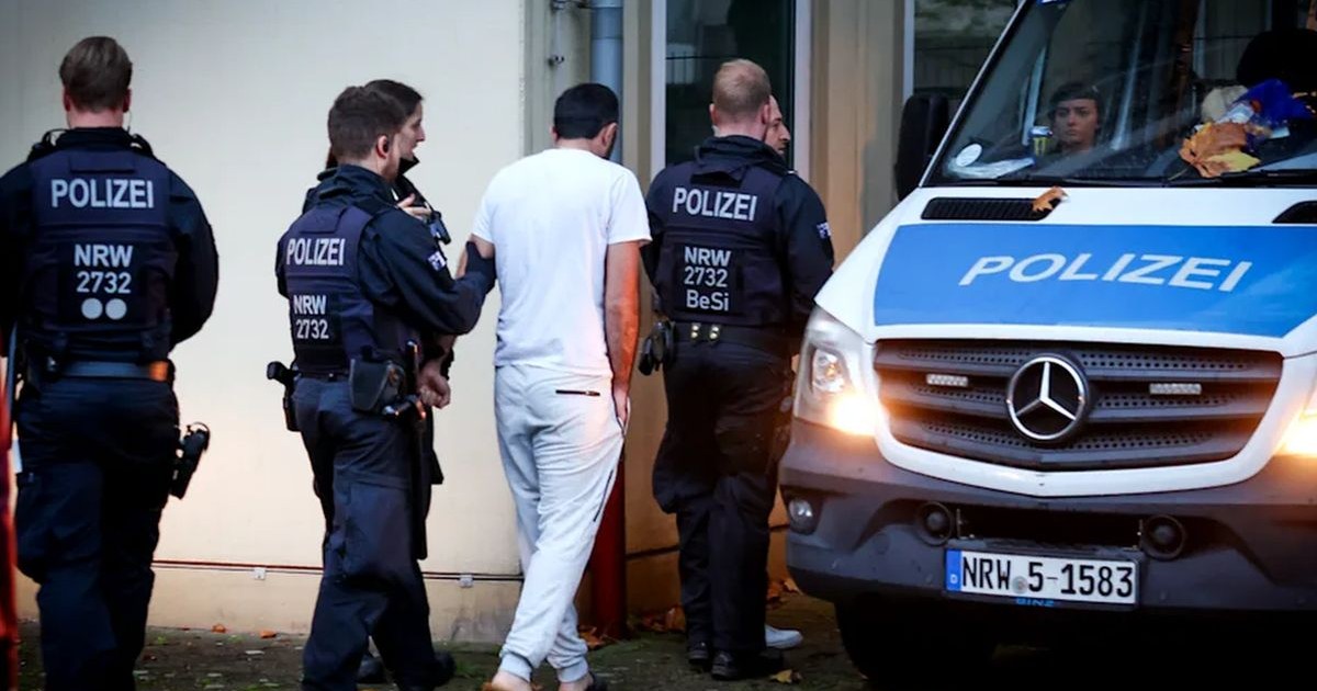 Un bărbat îmbrăcat în alb, cu spatele, este dus de poliția germană în sediul instituției, iar lângă ei se află autospeciala