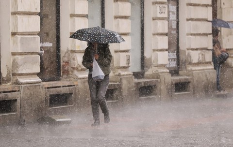 O persoană adăpostindu-se sub o umbrelă cu buline în timp ce merge pe trotuar pe o ploaie torențială, lângă fațada unei clădiri istorice.