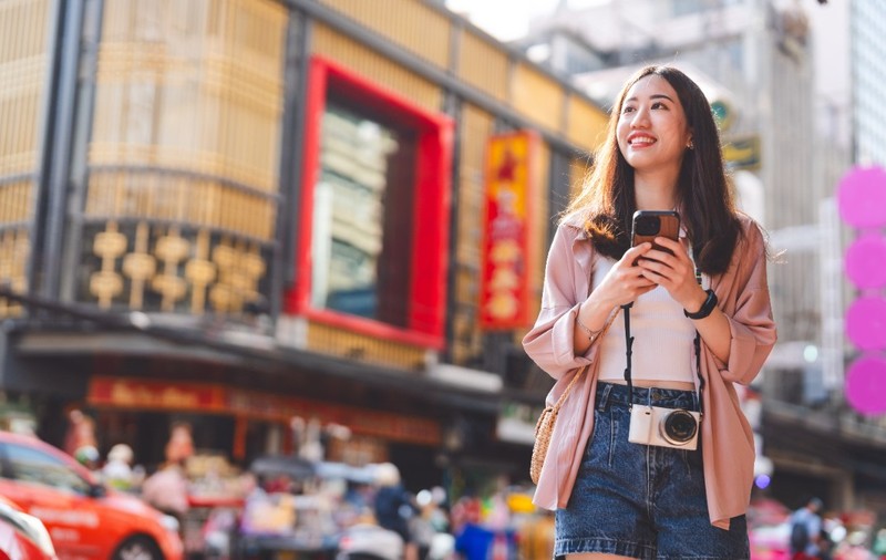 O femeie asiatică tânără cu un telefon în mână, pe o stradă din Hong Kong, în timp ce face o fotografie în fața unei clădiri înalte