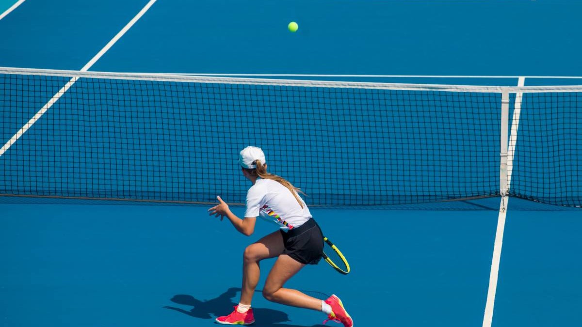 Jucătoare de tenis aflată în timpul unui meci pe un teren de tenis. Sportiva, îmbrăcată în tricou alb, fustă neagră și pantofi sport roz, poartă o șapcă albă și are părul prins într-o coadă.