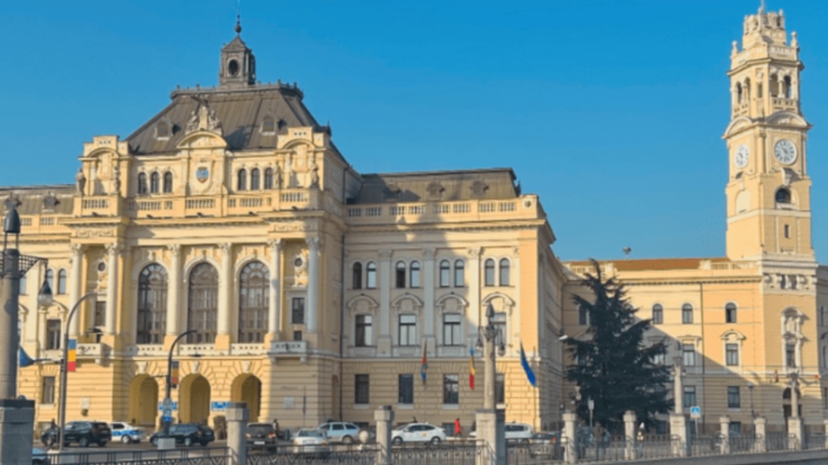 O clădire în nuanțe de galben pal/bej deschis. În partea dreaptă a clădirii este un turn înalt cu ceas