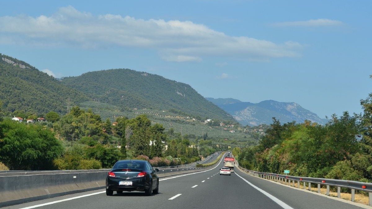 Imaginea prezintă un autoturism negru care circulă pe o autostradă ce șerpuiește printr-un peisaj cu dealuri împădurite, sub un cer senin de vară.
