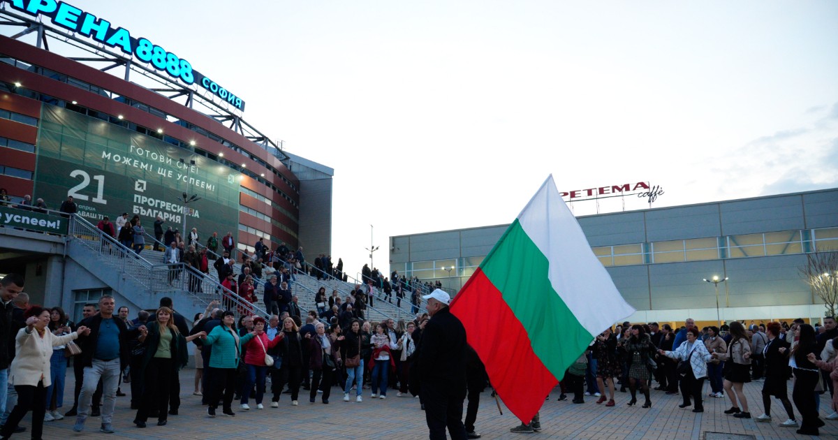 Un grup mare de oameni este adunat în fața unei clădiri din Sofia, participând la un eveniment public, în timp ce un bărbat ține în mână steagul Bulgariei. Atmosfera este una animată, specifică unui miting sau unei manifestații, cu mulțimea adunată în jurul scărilor.