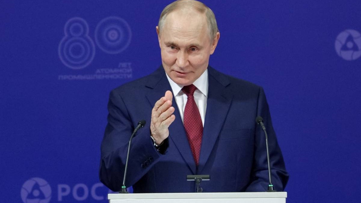 Vladimir Putin în timpul unei conferințe de presă, președintele rus arată cu mâna spre camera de fotografiat, îmbrăcat într-un sacou negru, o cămașă albă și o cravată roșie, stând în spatele unui pupitru alb pe care sunt montate două microfoane