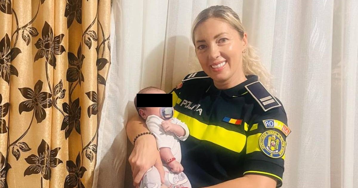 O polițistă blondă, îmbrăcată în uniformă, în timp ce ține un bebeluș în brațe și zâmbește în fața camerei.