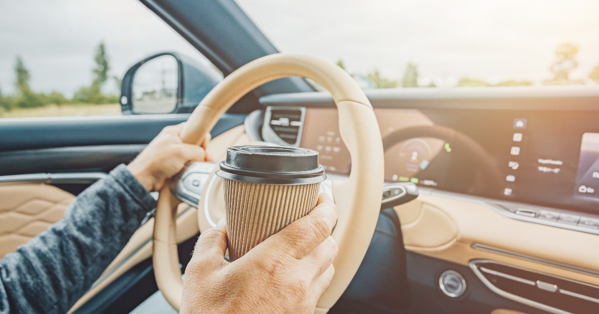 Un șofer ține o ceașcă de cafea în timp ce conduce, încercând să combată oboseala în timpul unei deplasări. Imaginea sugerează iluzia de vigilență oferită de cafea, care poate masca temporar epuizarea la volan.