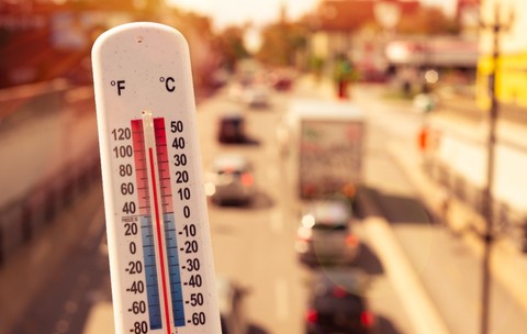 Prim-plan cu un termometru de exterior care indică o temperatură ridicată, pe fundalul unui drum urban însorit și aglomerat cu vehicule.