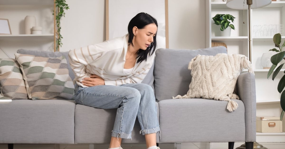 O femeie stă pe canapea și își ține mâna pe abdomen, vizibil afectată de un disconfort puternic. Expresia ei sugerează durere și îngrijorare, indicând o problemă de sănătate care poate trece ușor neobservată.