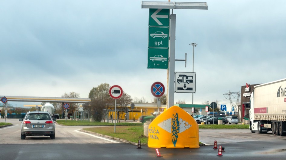 O zonă de servicii și stație de alimentare de pe o autostradă din Italia, cu panouri verzi de direcționare, indicatoare rutiere, un autoturism Volkswagen argintiu în mers și un camion staționat în dreapta.