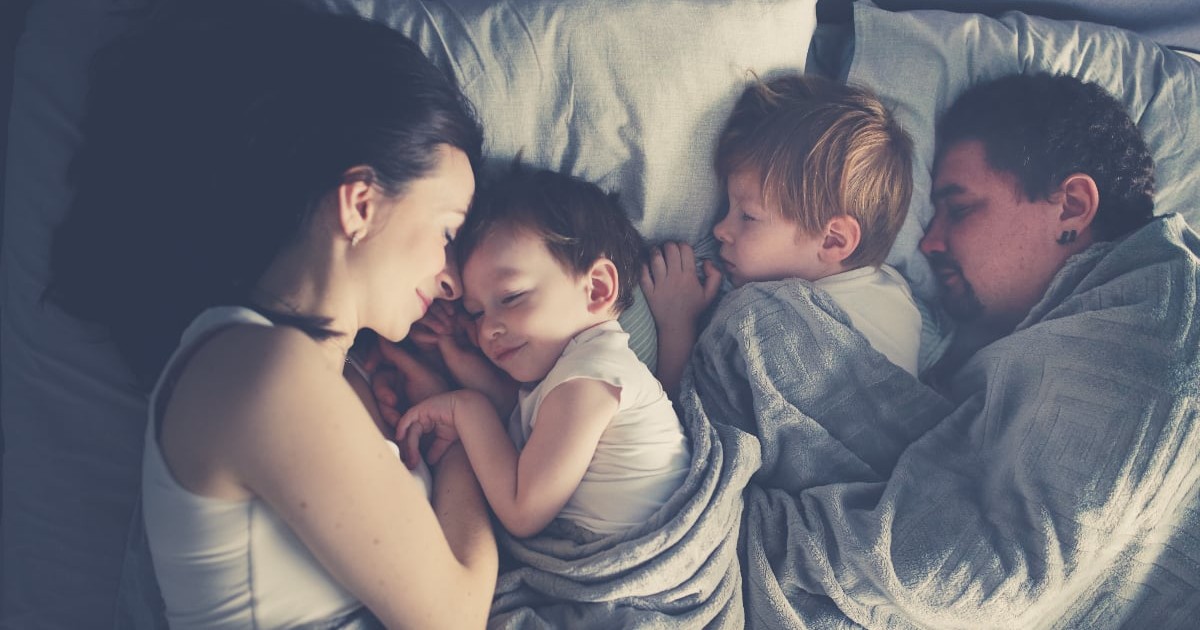 O familie doarme împreună în același pat, într-o atmosferă liniștită și caldă, sugerând apropiere și siguranță. Cei doi copii sunt între părinți, acoperiți cu o pătură, iar toți par relaxați și odihniți.