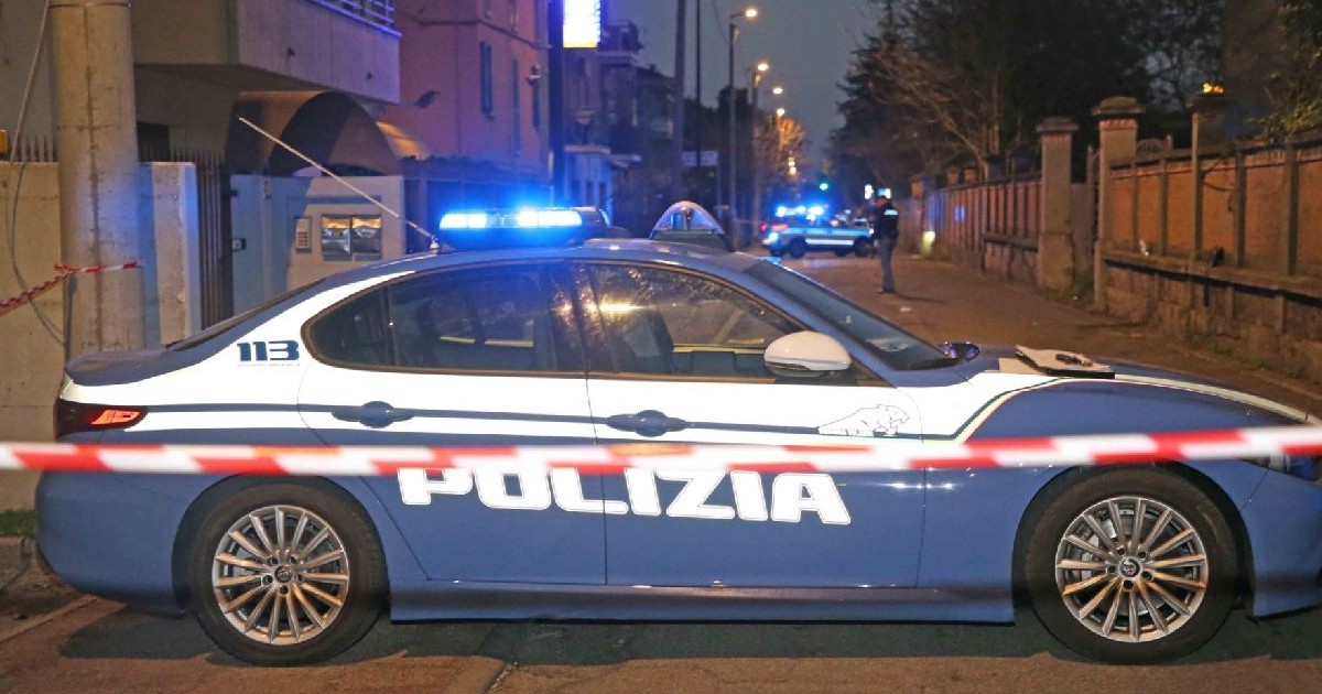 Fotografie cu o mașină de poliție italiană, cu lumini albastre intermitente, poziționată pe o stradă noaptea, izolată cu bandă roșie și albă.