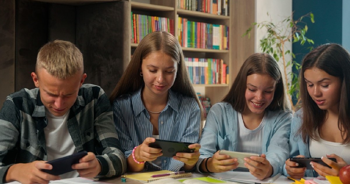 Patru elevi folosesc telefoane mobile, în timp ce sun așezați la o masă acoperită de cărți și caiete deschise, in spatele lor se vede o bibliotecă