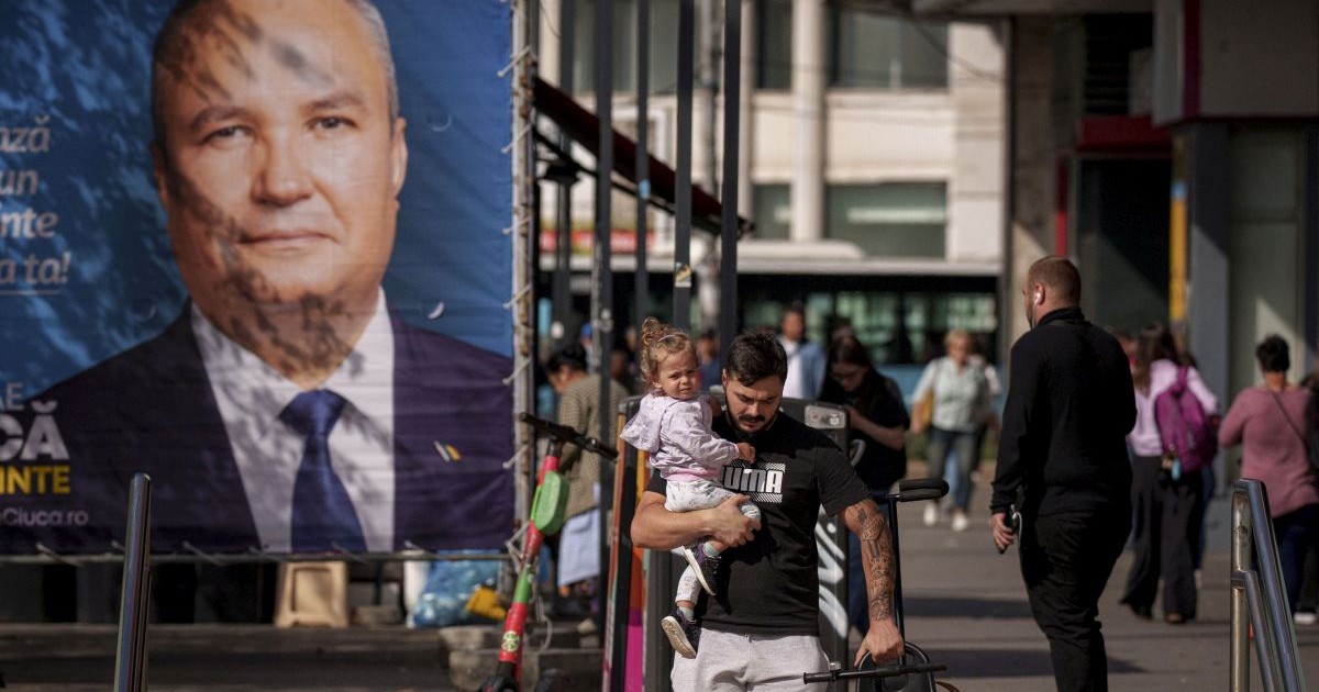 Un barbat care ține un copil în brațe trece prin fata unui afis electoral al lui Nicolae Ciuca, candidatul PNL pentru functia de presedintele al Romaniei, in Bucuresti, vineri, 20 septembrie 2024.