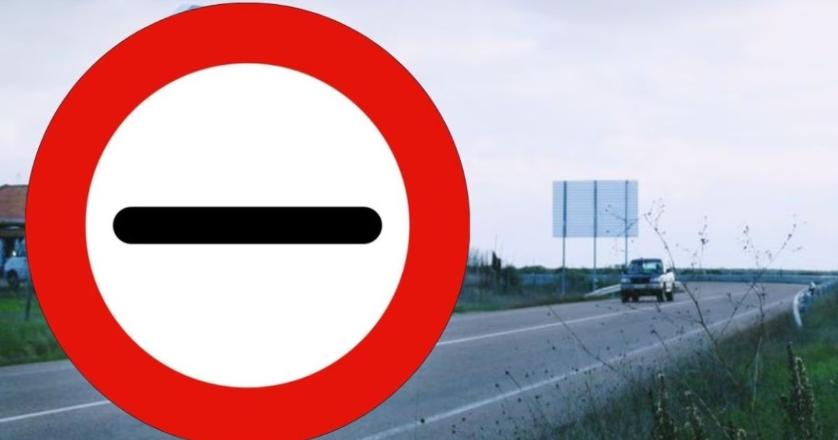 Imaginea prezintă în prim-plan un indicator rutier circular, alb cu margine roșie și o linie orizontală neagră în centru („Trecerea fără oprire interzisă”), amplasat pe marginea unui drum pe care se deplasează un vehicul.
