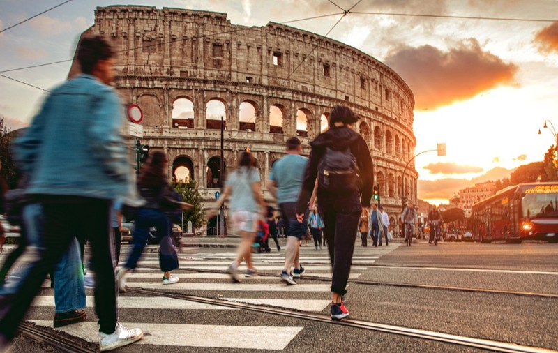 Imaginea prezintă pietoni traversând o stradă pe trecerea de pietoni în fața Colosseumului din Roma, pe fundalul unui apus de soare