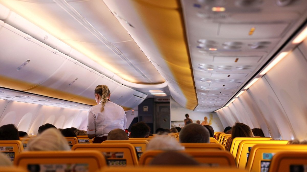Vedere a interiorului cabinei avionului din perspectiva pasagerului. Stewardesă elegantă, blondă, îmbrăcată în cămașă albă, care se ocupă de pasageri.