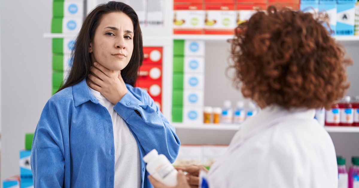 Femeie la farmacie, isi tine mana la gat in semn de durere. Farmacista este in fata ei si tine in mana o cutie de pastile