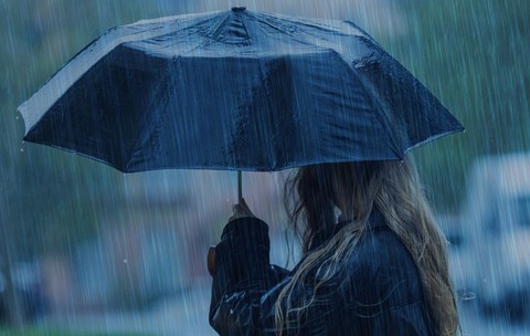 O persoană cu părul lung și blond, privită din lateral, care ține o umbrelă neagră deschisă pe o ploaie torențială.