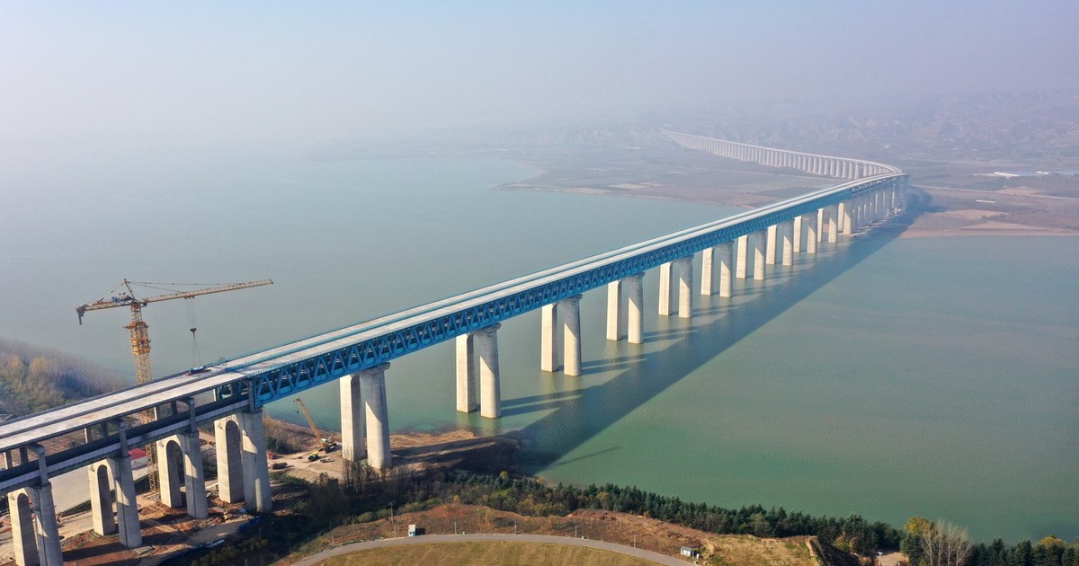 Această fotografie aeriană surprinde un viaduct feroviar monumental, cu piloni înalți de beton și o structură metalică albastră, care se întinde șerpuind peste un curs lat de apă și continuă spre orizont până se pierde în ceață, sub supravegherea unei macarale galbene de construcție.