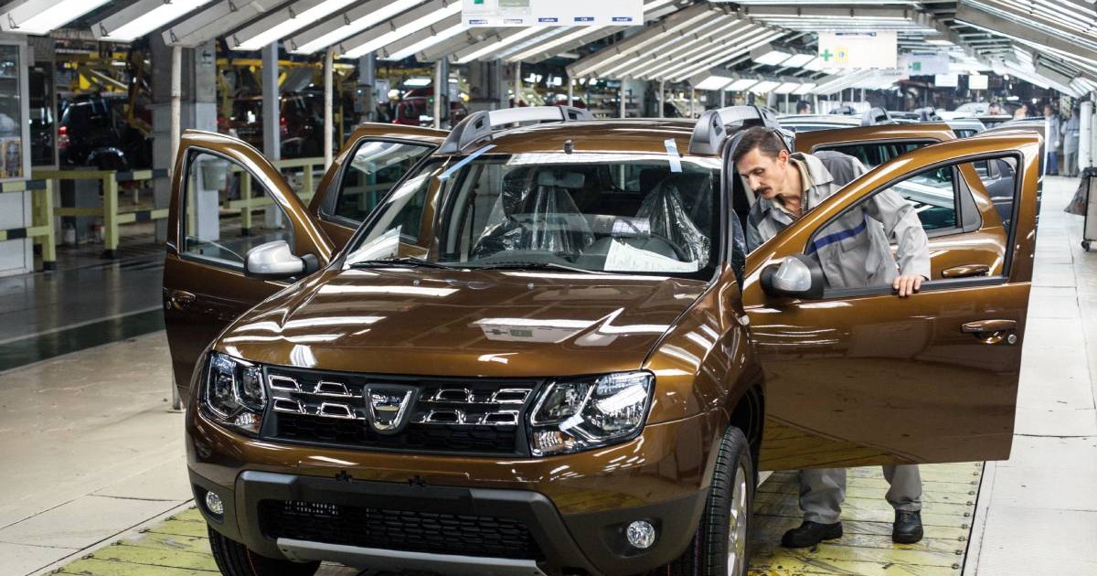 Interiorul unei fabrici auto Dacia din Mioveni, România. În prim-plan se află un model Dacia Duster de culoare maro aflat pe linia de asamblare, în timp ce un muncitor inspectează atent interiorul mașinii.