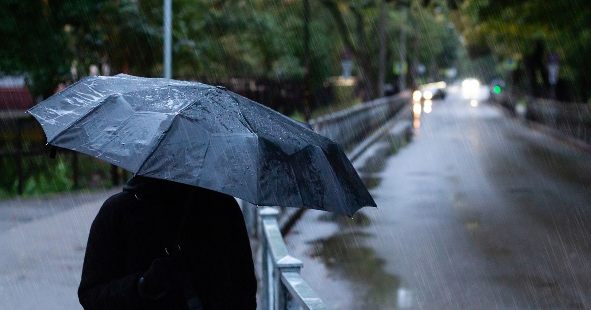 Un om îmbrăcat în negru și care are o umbrelă deschisă, fotografiat cu spatele, când merge pe stradă, într-o zi ploioasă și rece. În dreapta lui se vede o stradă pe care trec mașini