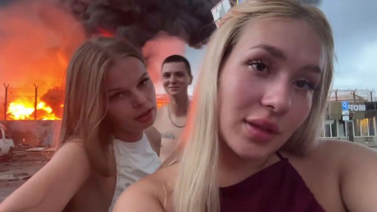Cei trei tineri din Rusia, în față două fete, una blondă și alta șatenă, iar pe fundal un tânăr și un depozit în flăcări,