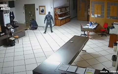 Secvență surprinsă de o cameră de supraveghere dintr-o încăpere, în care două persoane mascate și îmbrăcate în haine închise la culoare încearcă să spargă un seif metalic montat pe un perete.