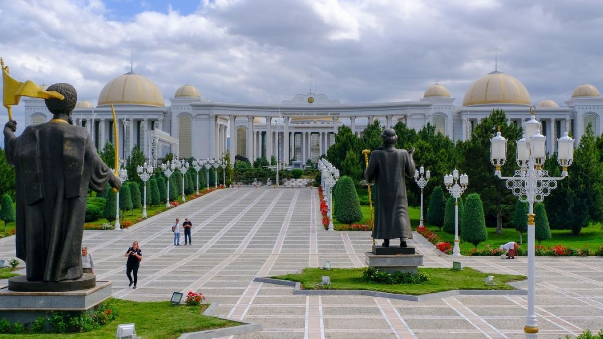 Biblioteca Națională din Așgabat, capitala Turkmenistanului Se vede o cladire impozanta, cu multe corpuri, alb[ cu acoperisuri aurite, alei largi pe care se plimba oameni, marginite de zone verzi, si doua statui care se vad din spate.
