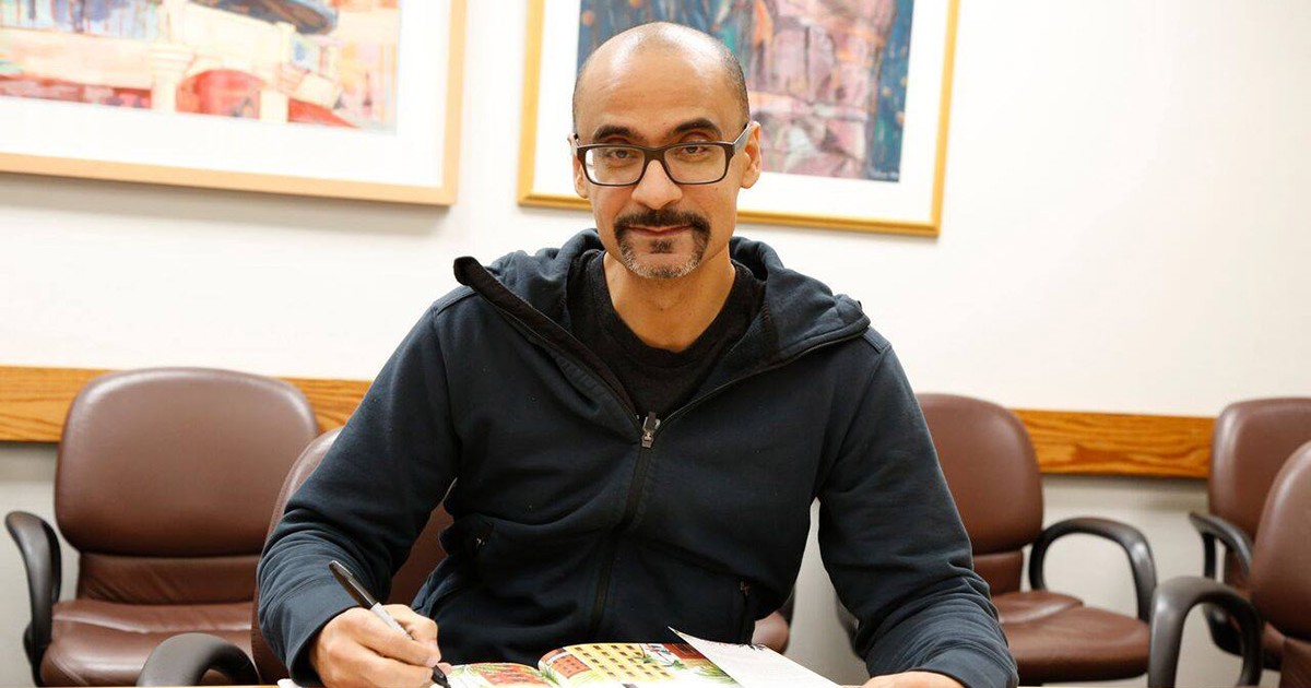 Junot Diaz, pe scaun la birou cu un pix in mâna dreaptă pe o carte deschisă