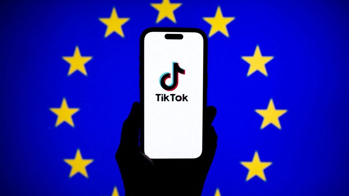 Logo-ul TikTok este afișat pe ecranul unui telefon mobil în fața unui ecran de computer care afișează steagul UE, ianuarie 2026