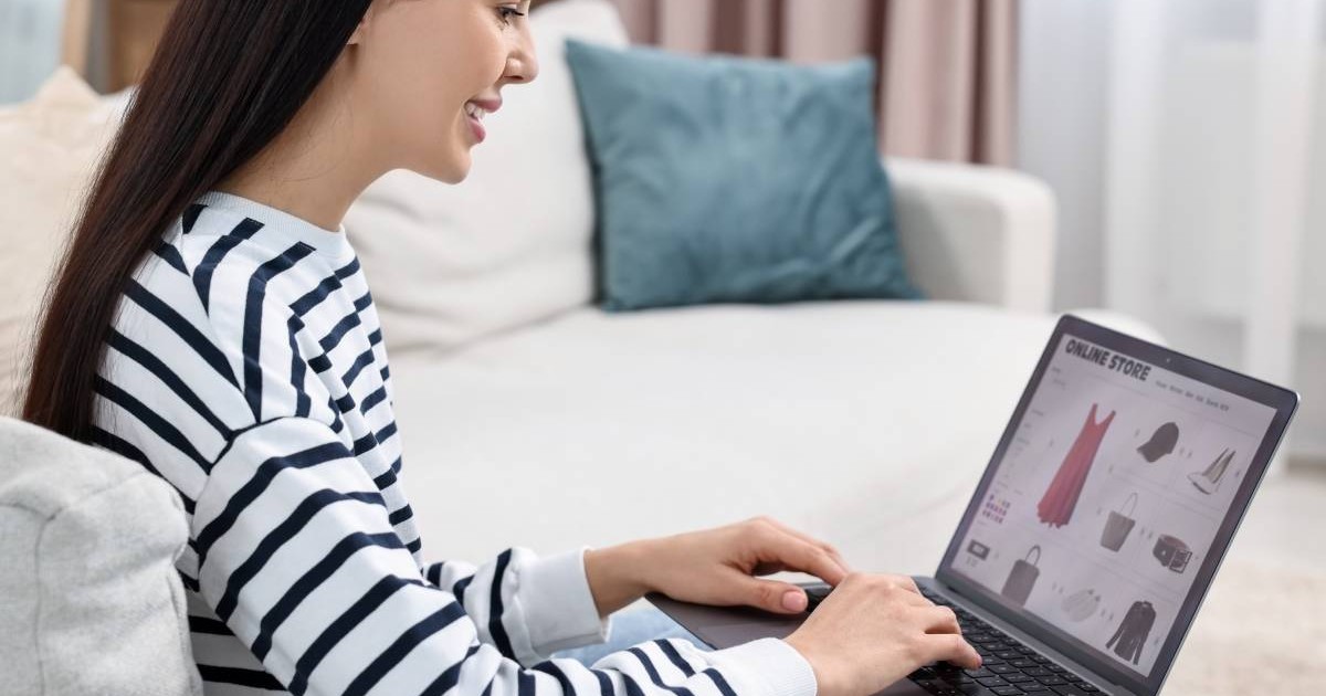 Femeie asezata, intr-o l=bluza alba cu dungi negre, cu un laptop in fata, face cumparaturi online