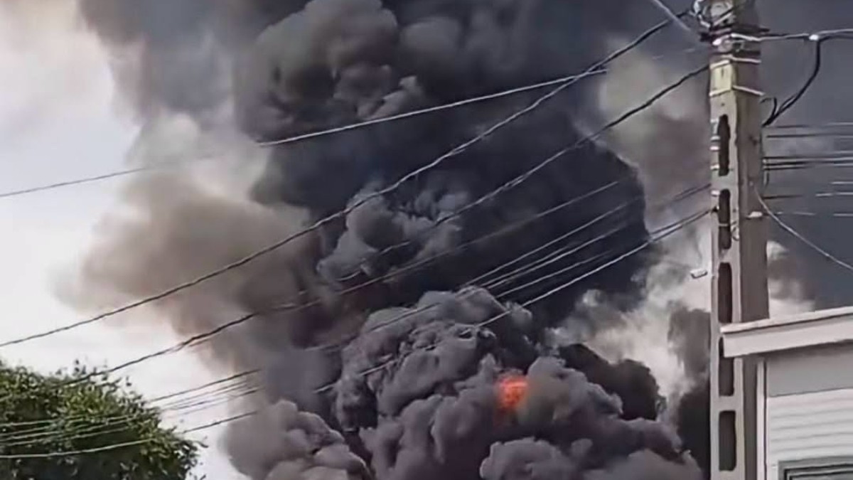 În imagine se observă un incendiu puternic, cu flăcări și un nor dens de fum negru care se ridică deasupra clădirilor. În prim-plan sunt vizibile mai multe cabluri electrice și un stâlp de electricitate.