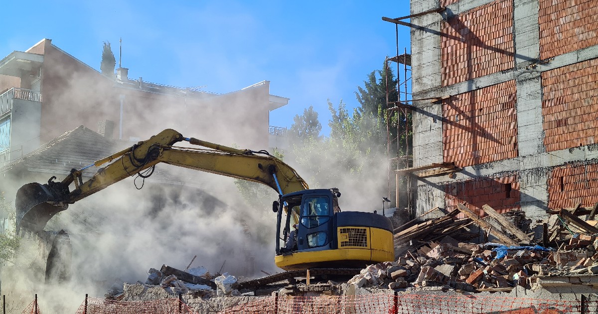 Un excavator demolează activ o structură, trimițând nori de praf în aer, pe fundalul clădirilor din apropiere