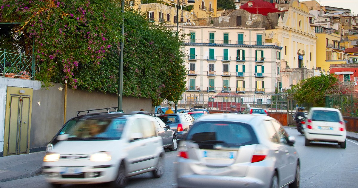 În imagine este prezentat traficul rutier aglomerat de pe o stradă urbană, cu mai multe mașini în mișcare ușor blurate, având pe fundal clădiri istorice în stil mediteranean specifice unui oraș din Italia.