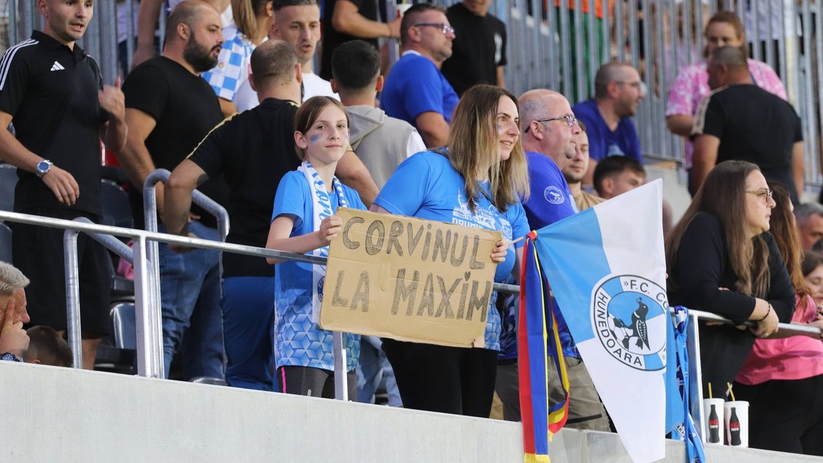 Suporteri ai echipei CorvinulHunedoara in Tribune. Majoritatea suporterilor poarta tricouri in culorile echipei, alb-albastru, iar pe o pancarta tinuta in mana d eo fetita se poate citi: Corvinul la maxim